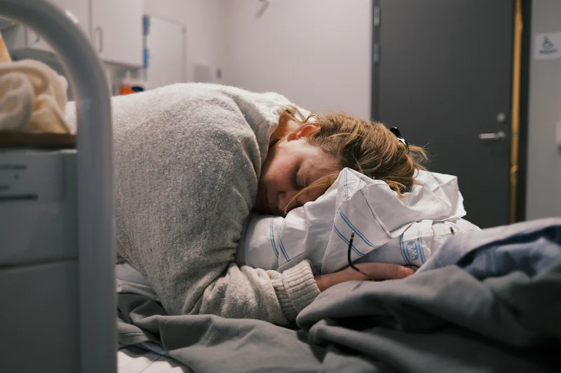 Woman lying in a hospital bed with an IV line and a blanket, eyes closed.
