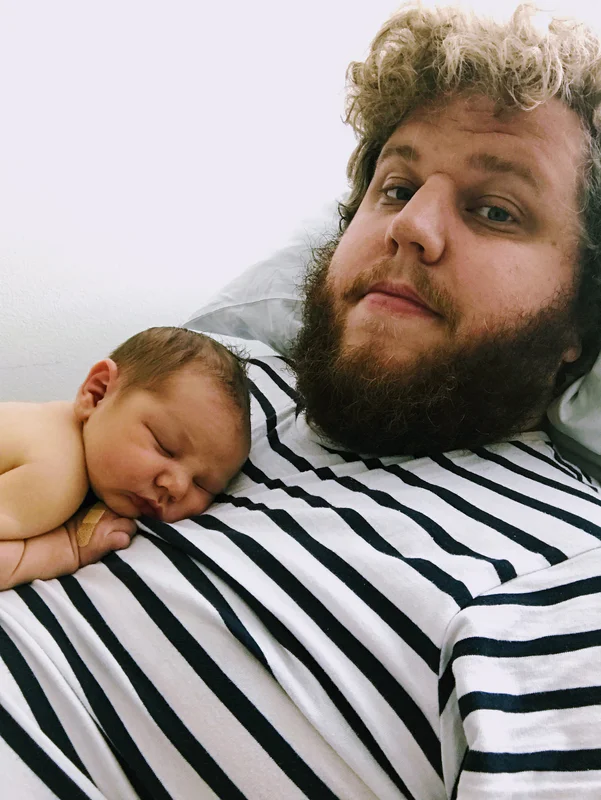 Bearded man wearing a striped shirt cuddles a sleeping baby in his arms against a plain background.