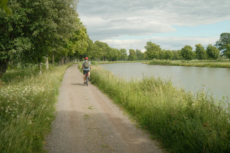 Person rides a bicycle on a gravel path beside a calm canal under a cloudy sky.