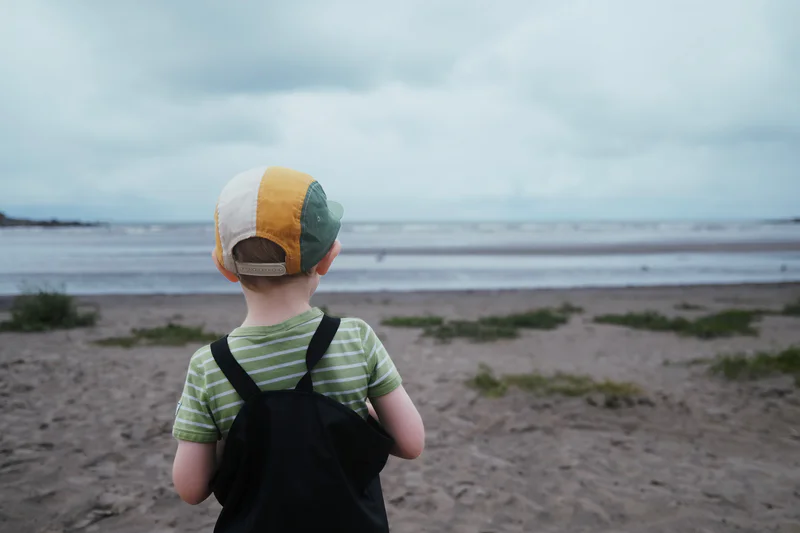 Child in green striped shirt and black overalls wearing a cap, standing on a beach facing the ocean.