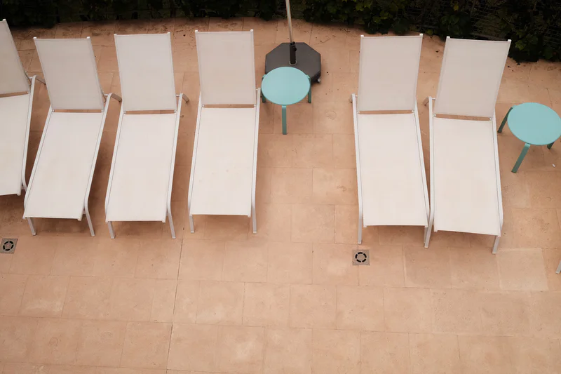 Six white lounge chairs lined up on a tiled patio beside two round teal tables.
