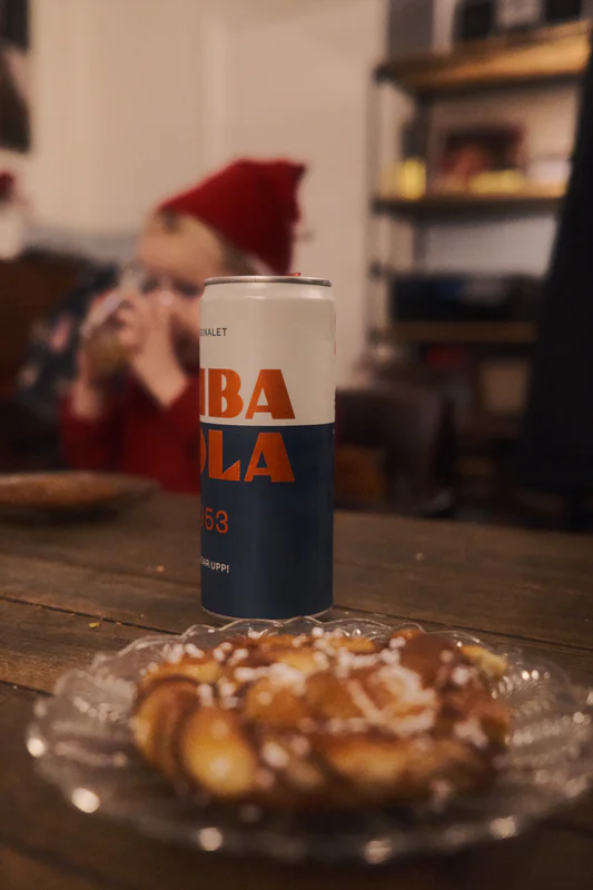 Crisp can of “Cuba Cola” on a wooden table beside a glass plate with a bun; blurred people and shelves behind.