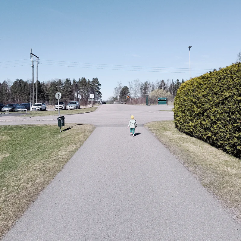 Person walking away down a paved path beside parked cars, hedges, and trees under a clear sky.