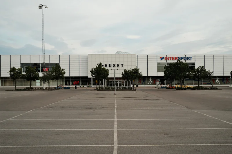 Large shopping center façade with I-HUSET and INTERSPORT signs, trees, and an empty parking lot.