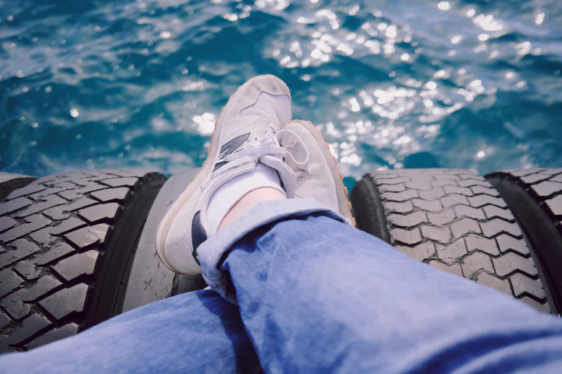 Person’s feet in sneakers stretched on a dock over rippling ocean water, with black tire tread in the foreground.