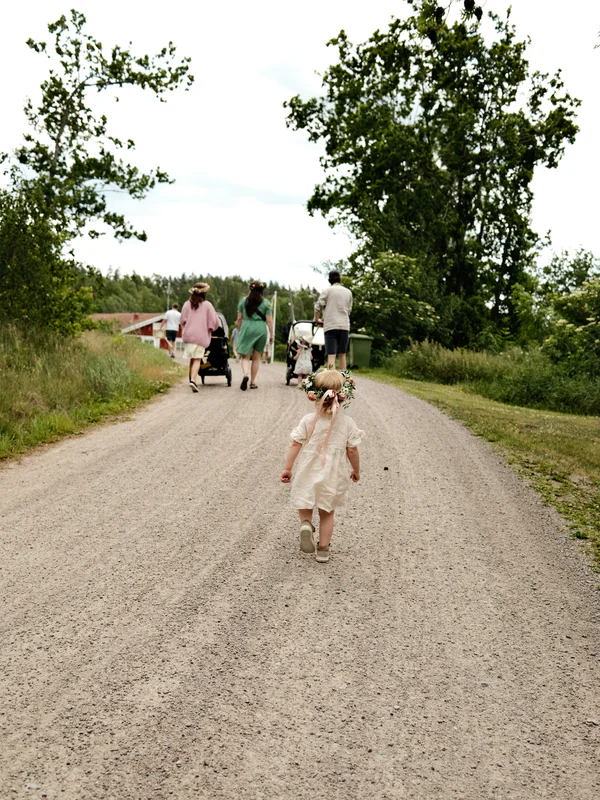 People walking along a gravel path through greenery, including a child carrying a small flower crown.
