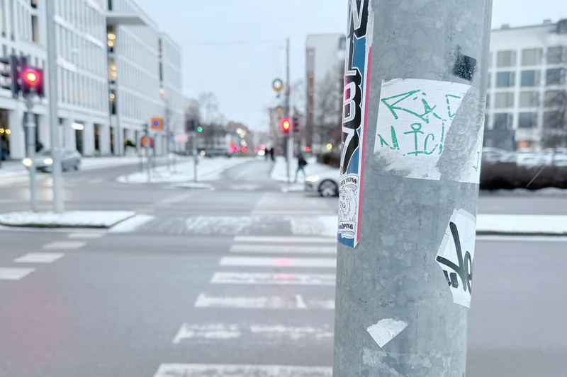 Street intersection with snow and a concrete pole covered in stickers, graffiti, and tape in foreground.