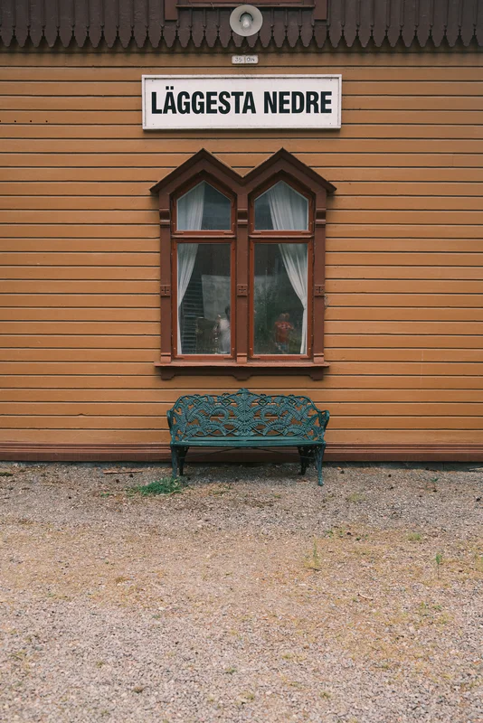 Teal decorative bench in front of a yellow wooden wall with a window and sign reading “LÄGGESTA NEDRE.”