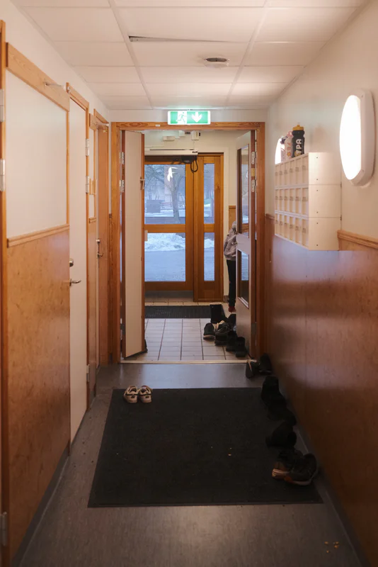 Narrow hallway with wooden doors, wall lockers, shoe mats, and a closed door at the far end.