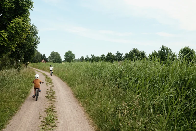Two cyclists ride along a narrow dirt path beside tall green grass under a cloudy sky.