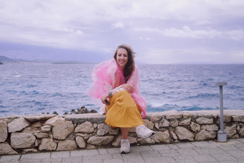 Woman smiling in a pink sheer poncho and yellow skirt, posing by a stone wall overlooking the ocean.