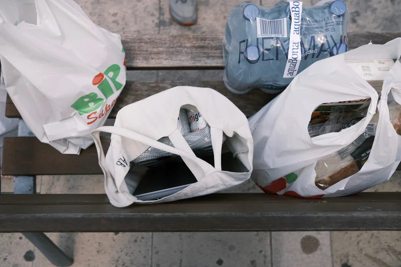 Assorted packaged items in white plastic bags and drink bottles on a bench in a street setting.