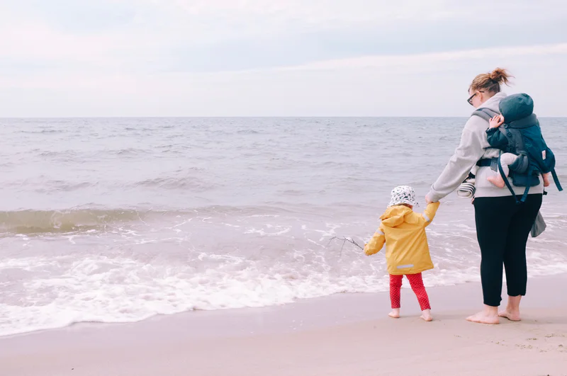 A woman holds a child at the shoreline as waves roll in under a cloudy sky.