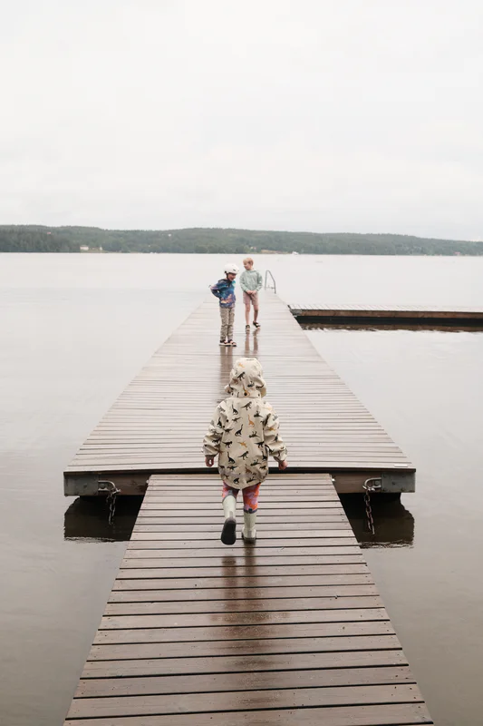 Three children stand on a wooden pier over calm water, with a lake and distant trees in the background.