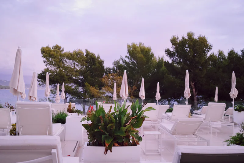 Row of white patio chairs with folded umbrellas and flower arrangements on a terrace with trees in the background.
