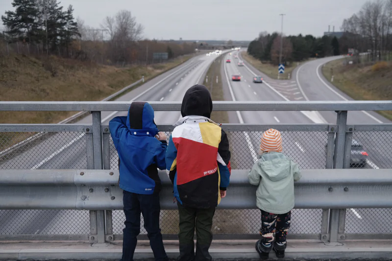 Two adults and a child watch cars on a highway from behind a roadside metal fence, cloudy sky.