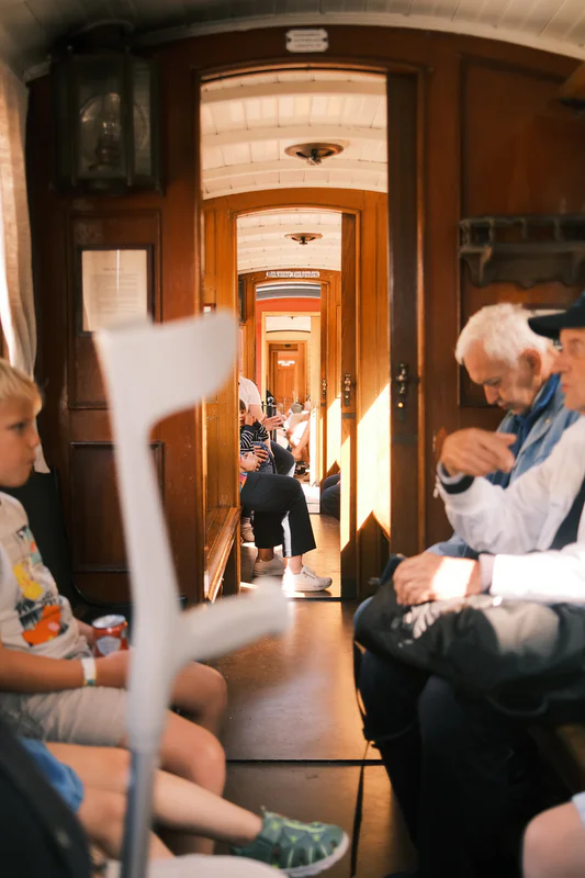 View down a train carriage aisle with passengers seated on both sides and people standing in the distance.