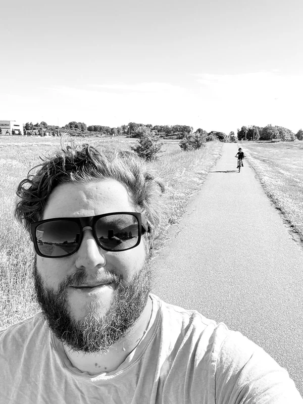 Man wearing sunglasses takes a selfie on a rural road through grassy fields with trees in the distance.