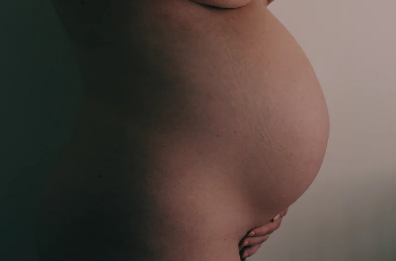 Close-up of a bare pregnant belly and arm resting at the hip against a plain background.