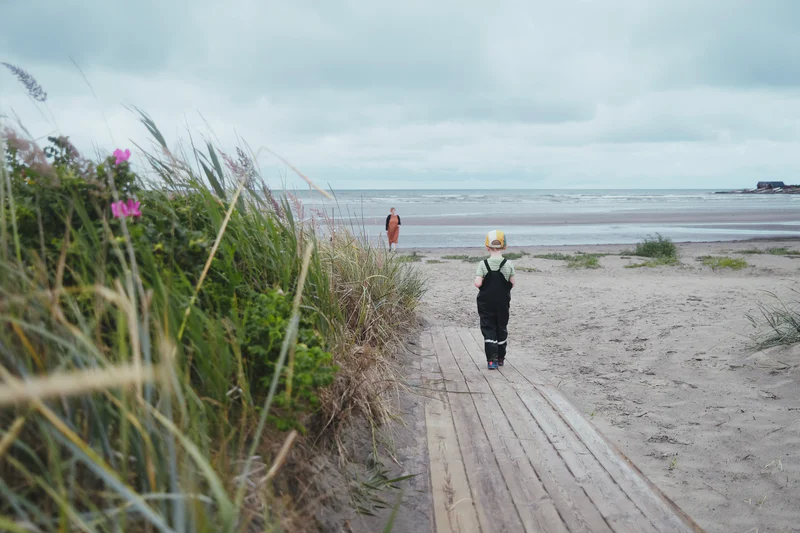 Two people walk along a sandy beach path bordered by tall grass toward the ocean under cloudy skies.
