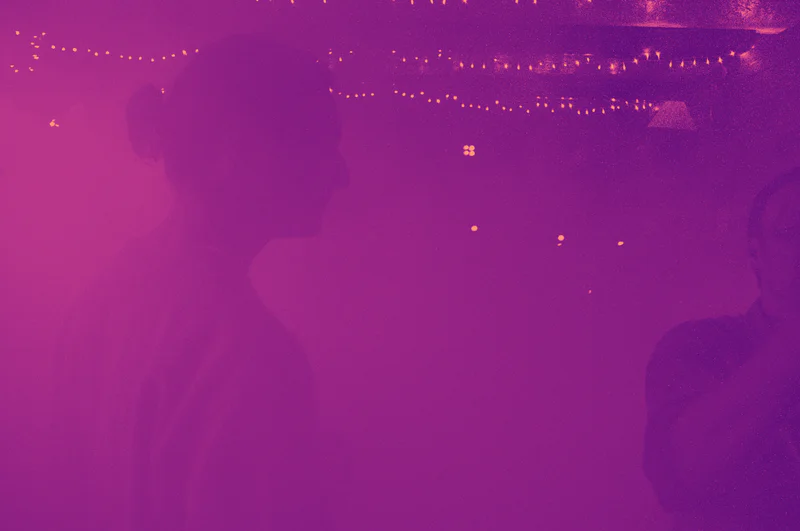 Purple-tinted scene with a dark silhouette of people and strings of small lights overhead.