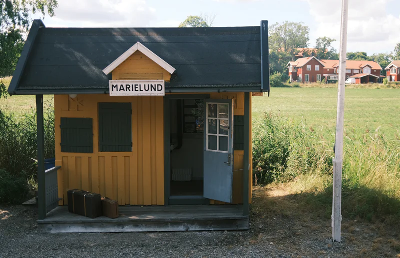Small kiosk with a “MARIELUND” sign, green doors and windows, set in a grassy field near houses.