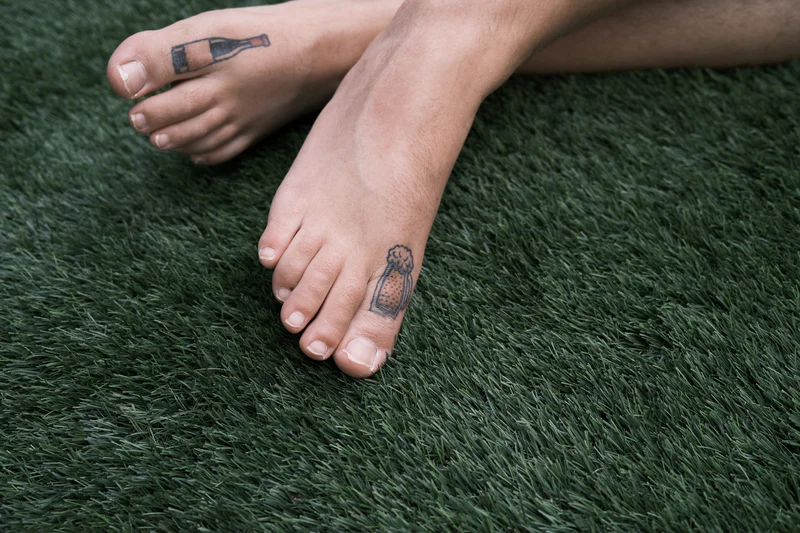 Two bare feet on green grass with small bottle-shaped tattoos on the toes.