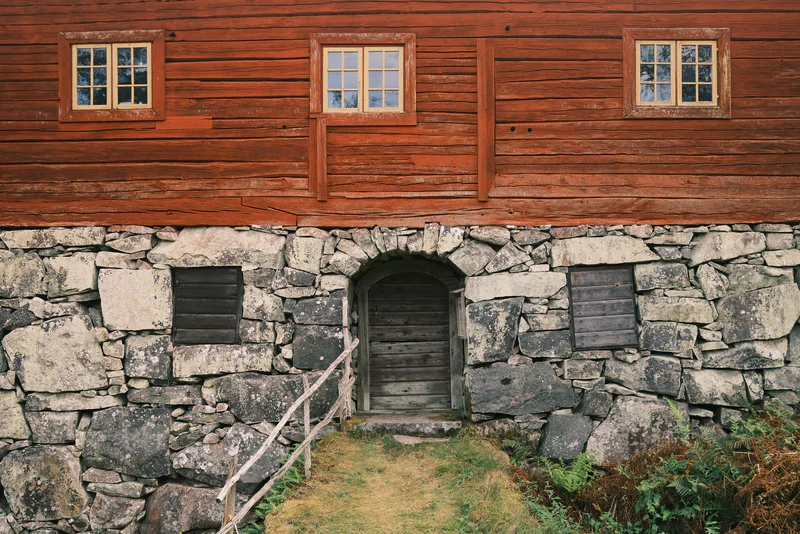 Red wooden house with three windows above a gray stone foundation and central arched doorway.