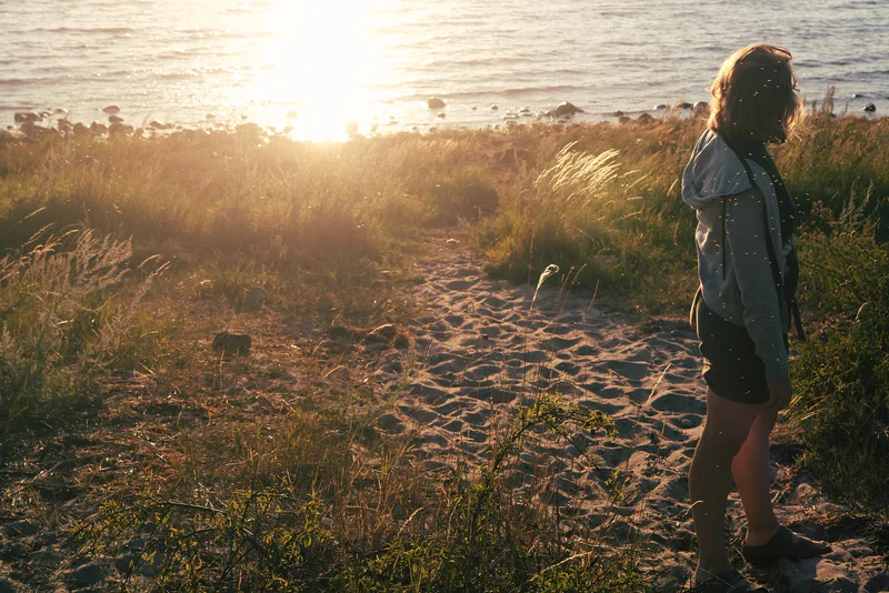 Person standing on a sandy beach along tall grass at sunset, facing the ocean.