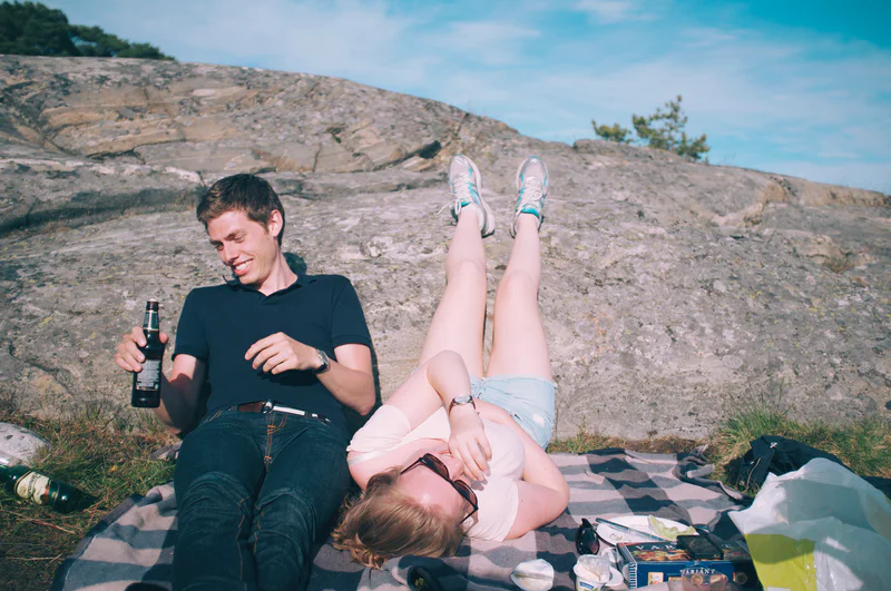 Man sits on a blanket beside a reclining woman holding a wine bottle, with rocky hills in the background.