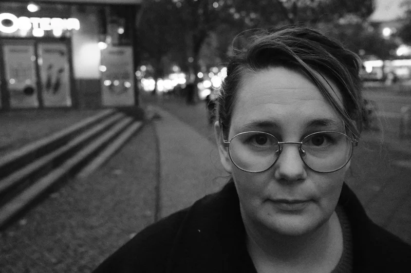 Person with round glasses and dark coat outdoors at night, street lights and buildings in background.