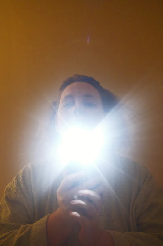 Woman wearing a dark robe holds up a bright phone flashlight, with face illuminated against a plain wall.