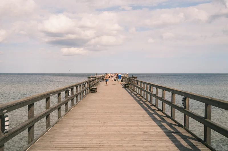 Wooden boardwalk over the ocean with people walking between two railings under a cloudy sky.