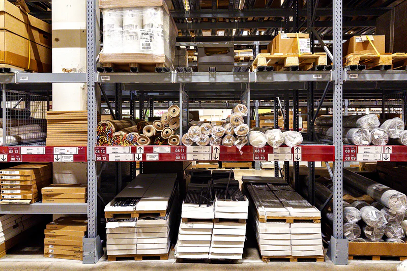 Warehouse shelves with boxed supplies, stacked pallets, and bundled materials on metal racks.