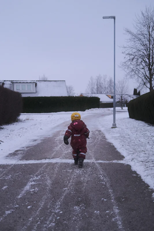 Child in a red winter snowsuit with a yellow hat walks on a snowy driveway beside hedges and a streetlamp.
