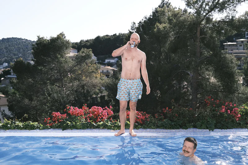 Shirtless man next to a pool drinking from a beer can while another man smiles from the water.