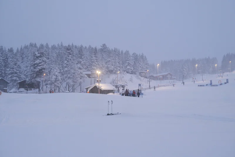 Snowy ski slope with lights and buildings in a forested valley under dusk foggy sky.