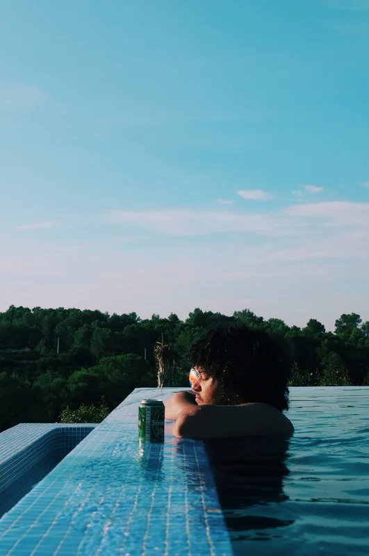 Person in a pool holding a can, seen from above against a background of trees and sunset sky.