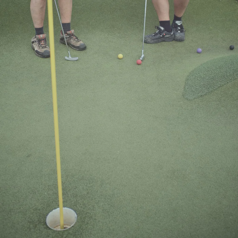 Two players’ legs in socks stand at a mini-golf green with a putter near colorful balls.