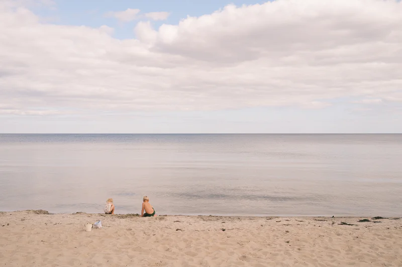 Two people sit on a sandy beach near the shoreline with calm ocean and a cloudy sky.