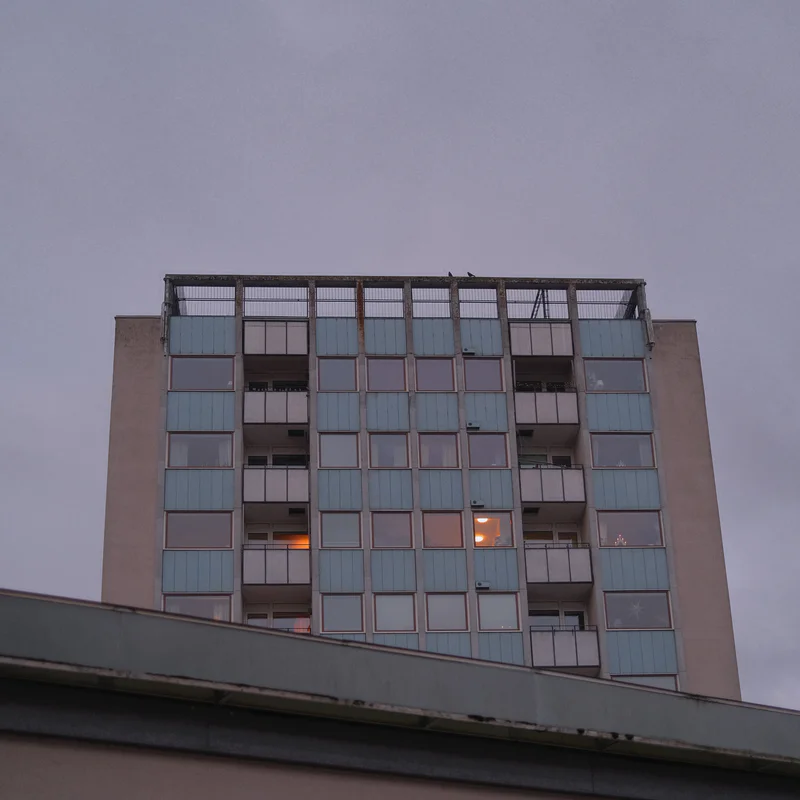 Tall apartment building with many windows at dusk, seen from a low angle against a gray sky.