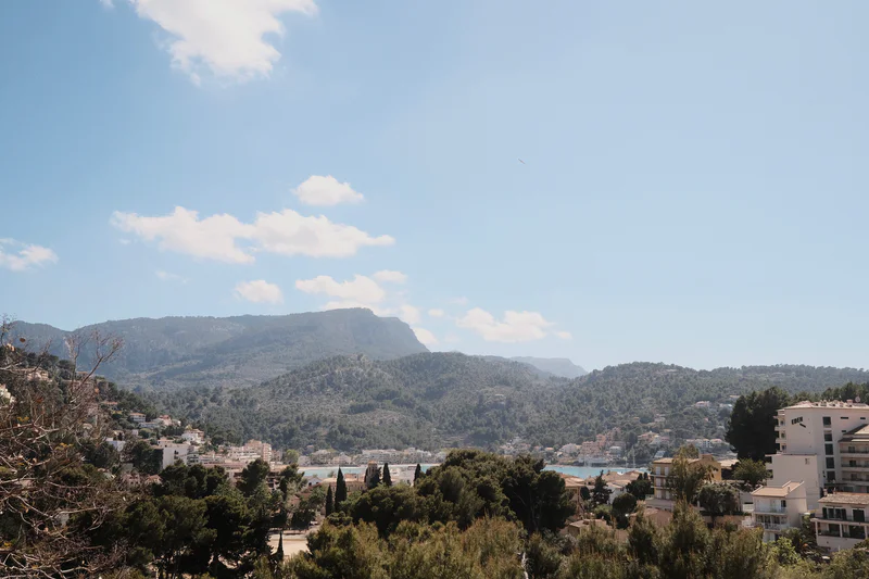 Mountain landscape with houses and trees in the foreground under a blue sky with scattered clouds.