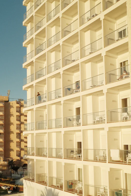 Sunlit hotel building façade with stacked balconies and many windows against a blue sky.