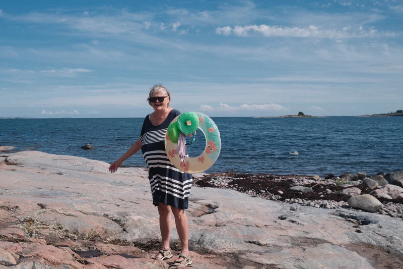 Smiling woman in a striped dress stands on rocky shore holding a colorful inflatable ring by the sea.