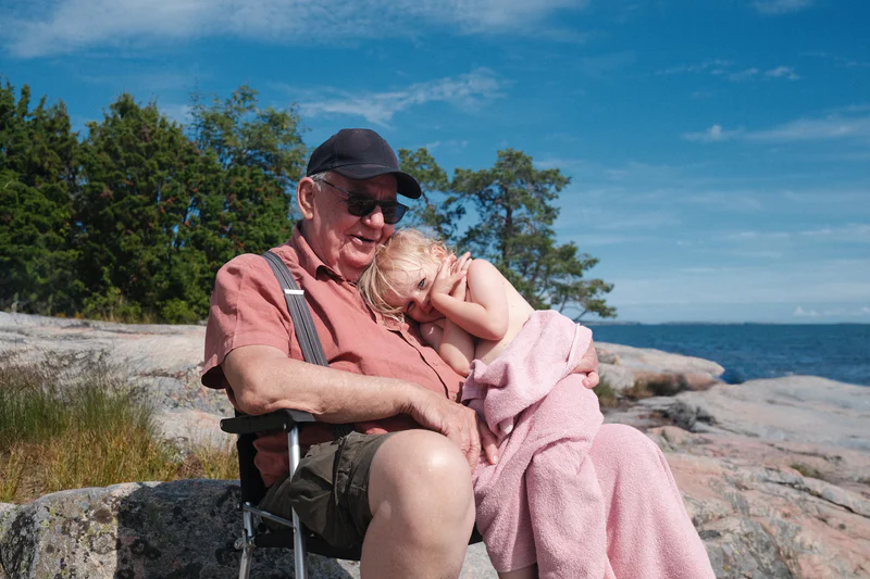 Man in cap and sunglasses holds a smiling blonde child wrapped in a pink towel by the ocean shore.