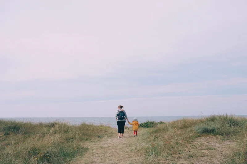 Two people walk along a grassy coastal path toward the ocean under an overcast sky.