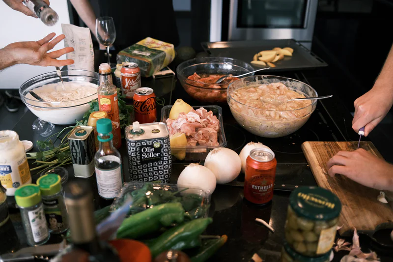 Cooking ingredients and spices laid out on a counter, with bowls of meat and dough and people preparing food.