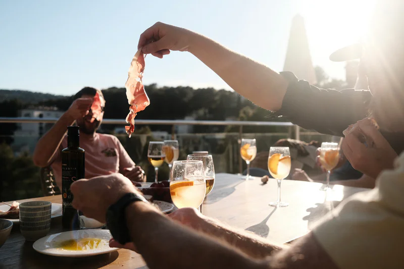 People on a terrace hold slices of ham over glasses of white wine in bright sunset light.
