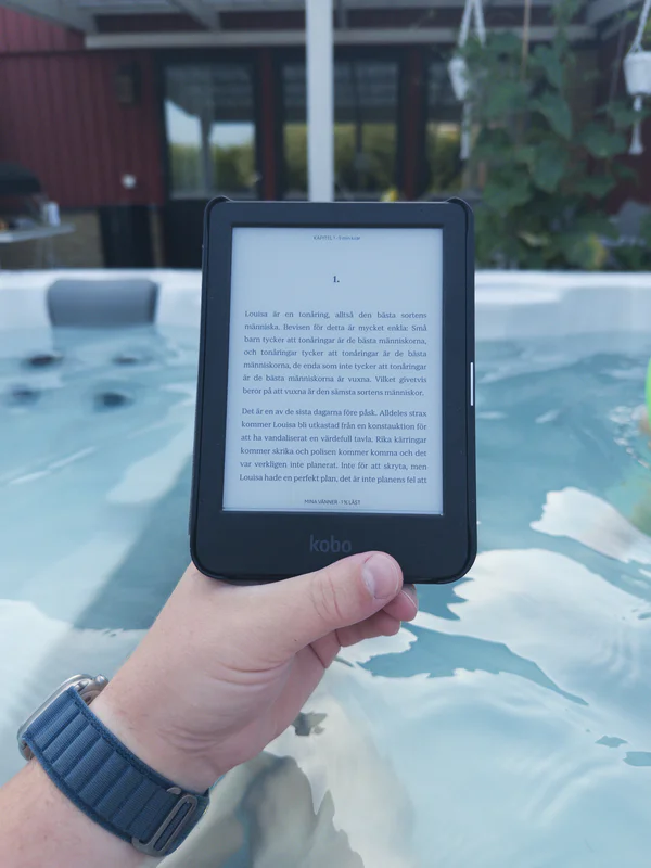 Hand holding a Kobo e-reader displaying Swedish text outdoors by a pool, with a building and trees in background.