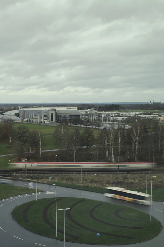 Blurry train passes along a highway beside industrial buildings under cloudy skies.